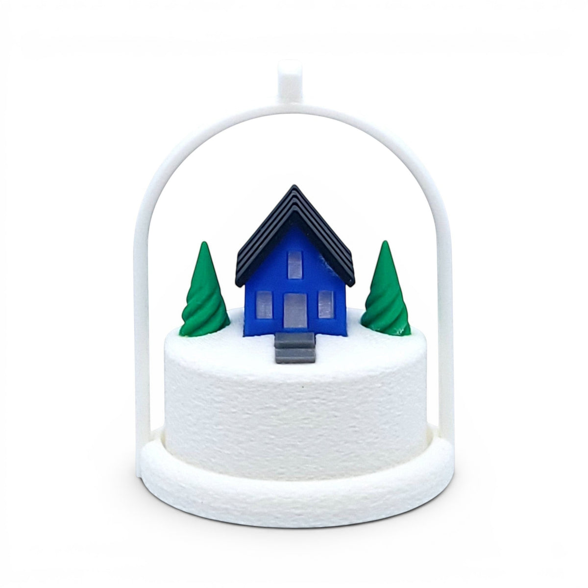 Miniature blue house ornament with green trees on a snowy base inside a white arched frame. Front view of blue house, in tree stand with light off.