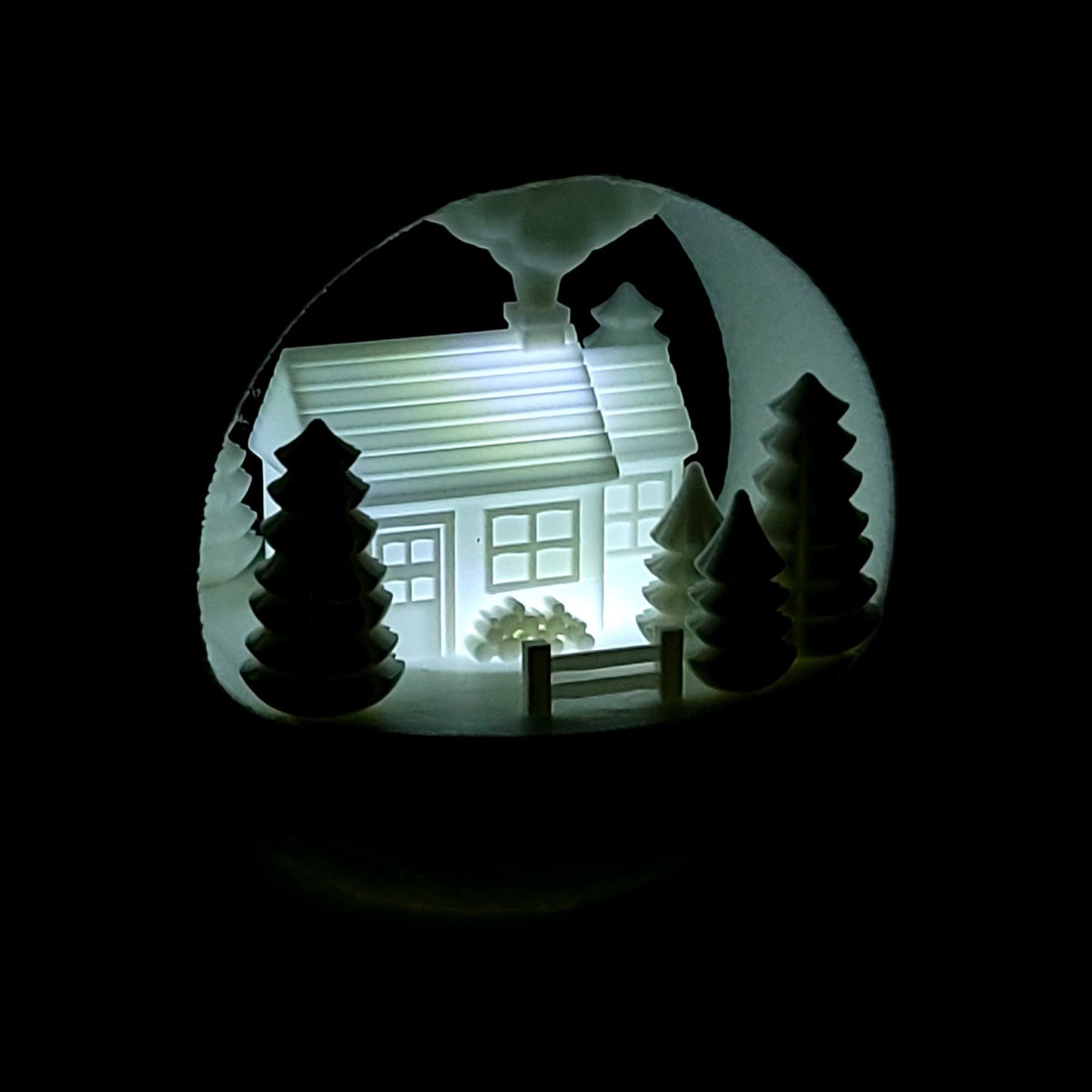 illuminated winter farmhouse ornament with evergreen trees and fence, made of PLA plastic. Light on, dark background, front of ornament.