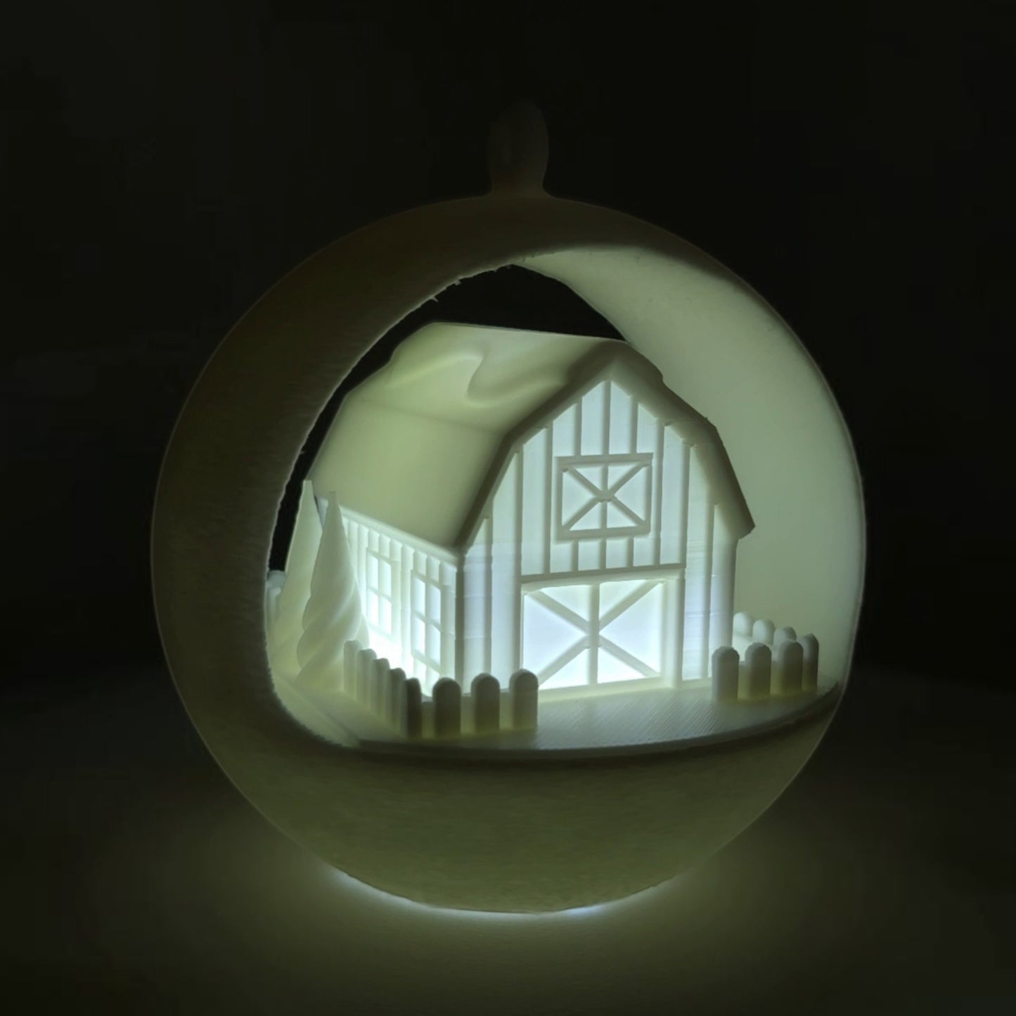 illuminated winter barn ornament with picket fence and trees, made of PLA plastic. Light on, rear left corner of ornament, background lights off.