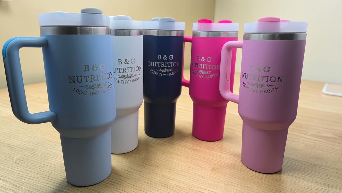 Five insulated travel tumblers with handles are lined up on a beige surface against a textured metallic background. The tumblers come in light blue, white, dark navy, bright pink, and pastel pink, each printed with the text 'B&G NUTRITION HEALTHY HABITS'.