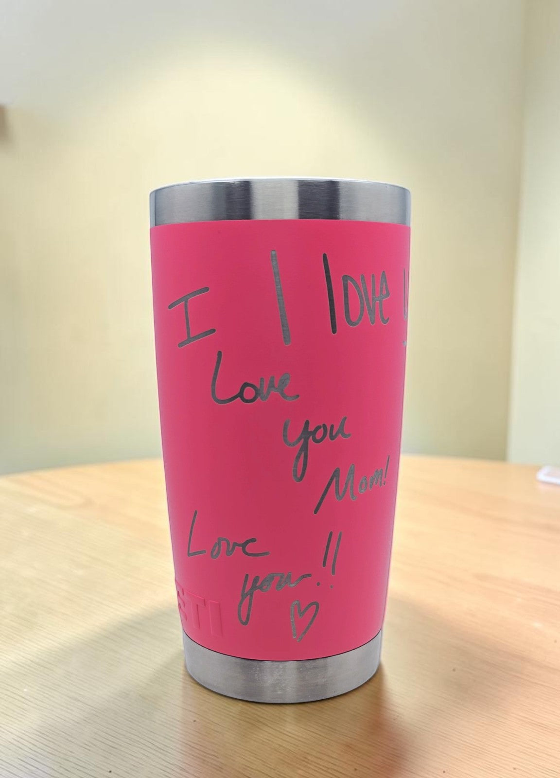 A pink insulated tumbler with a stainless steel rim and base, featuring handwritten laser engraved text that reads 'I LOVE Love you Mama! Love you!!' on a wood table.