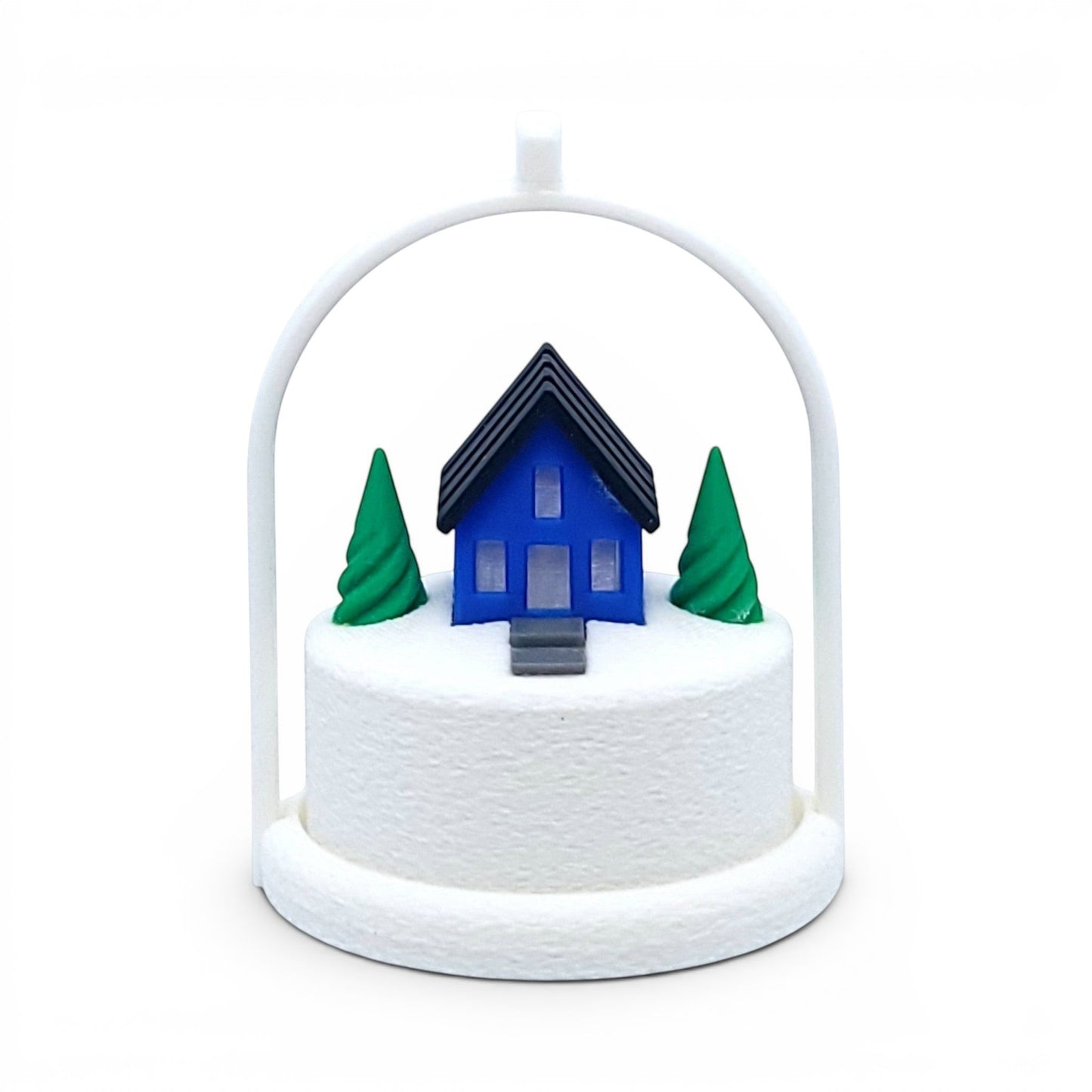 Miniature blue house ornament with green trees on a snowy base inside a white arched frame. Front view of blue house, in tree stand with light off.