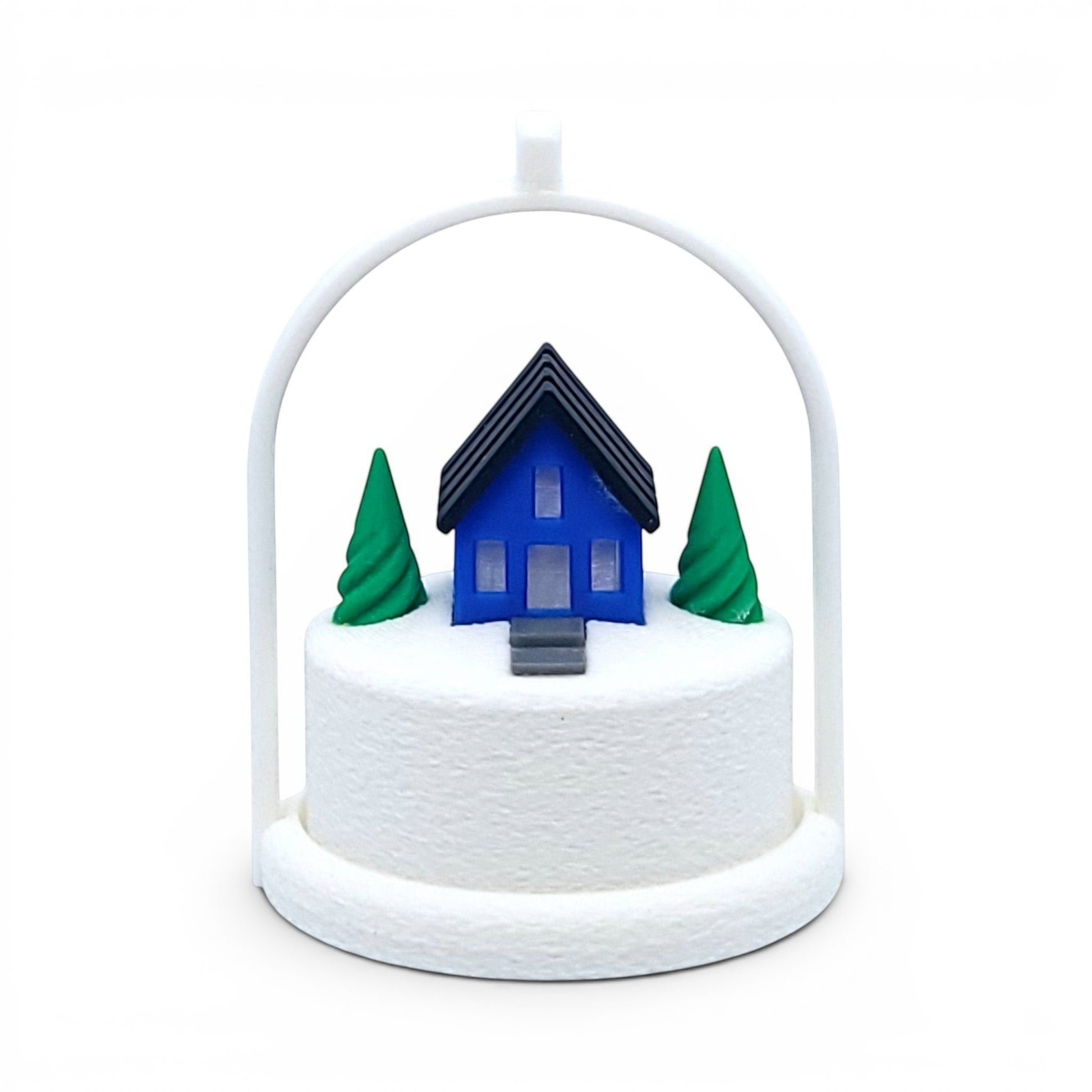 Miniature blue house ornament with green trees on a snowy base inside a white arched frame. Front view of blue house, in tree stand with light off.