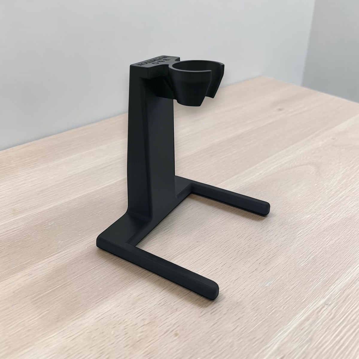 A matte black 3D-printed immersion blender stand with a vertical post, angled support arms, and a top cup holder, shown on a light wood tabletop against a neutral gray wall.