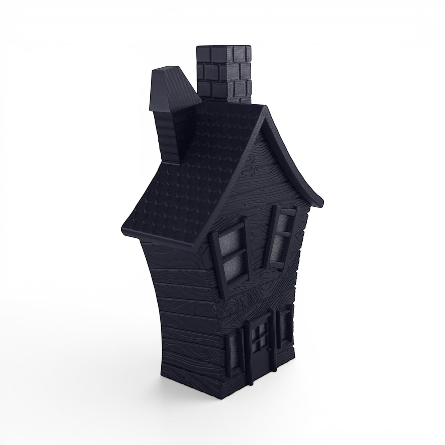 Black PLA flickering haunted house decoration with textured wood grain finish, crooked chimney, and window details. Front left corner of house.