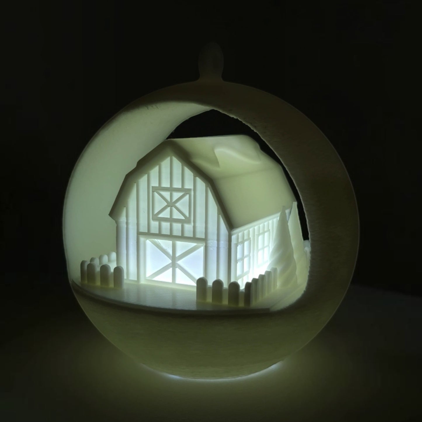 illuminated white barn ornament with picket fence and trees, PLA plastic, holiday decor. Light on, front right corner of ornament, background lights off.
