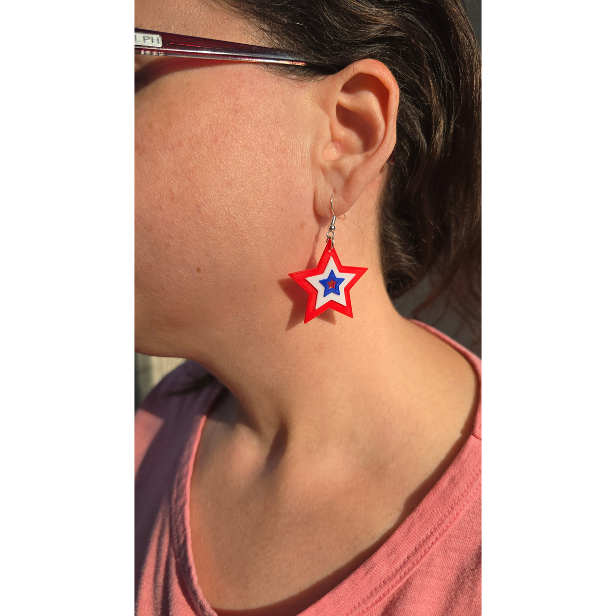 layered star dangle earrings in red, white, and blue worn by a woman in a pink shirt.