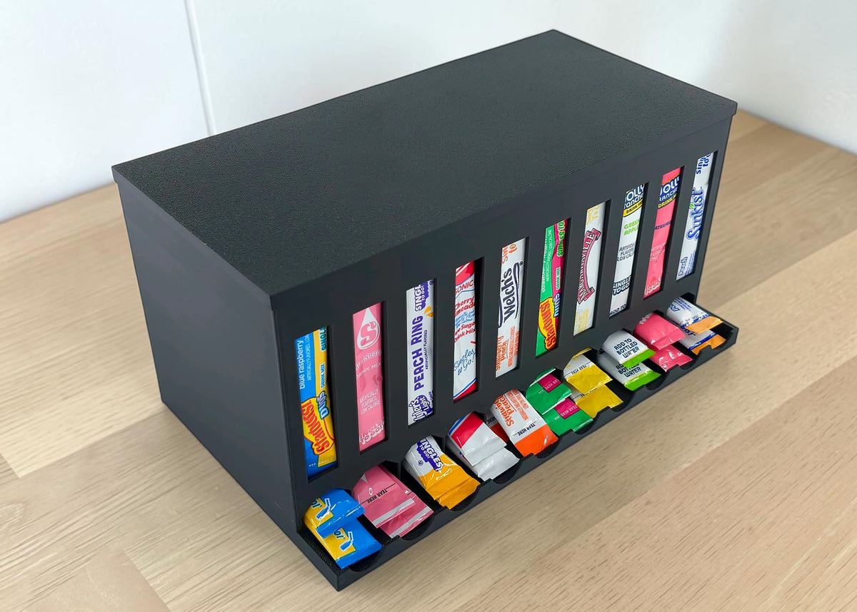 A black rectangular powder drink mix dispenser box with multiple vertical slots, placed on a light wood tabletop against a white wall.