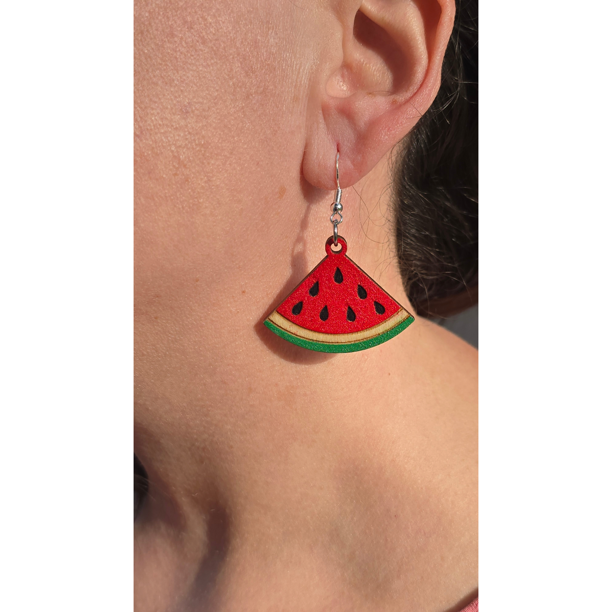 watermelon dangle earrings made of hand-painted basswood with hypoallergenic silver hooks hanging from ear for scale.